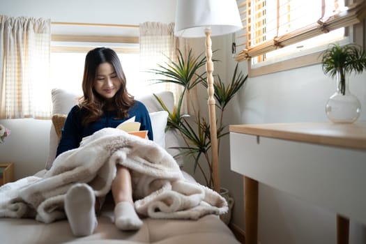 Smiling young woman reading a book by the Window, wrapped in soft blanket.