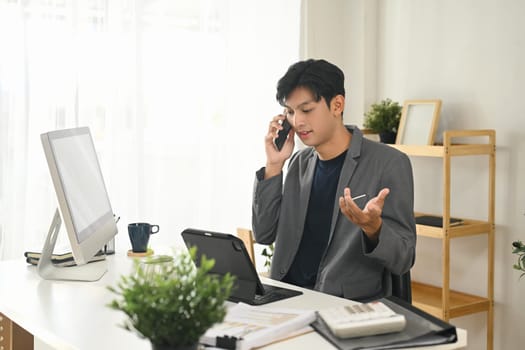 Young businessman in suit having phone conversation and using digital tablet at office.