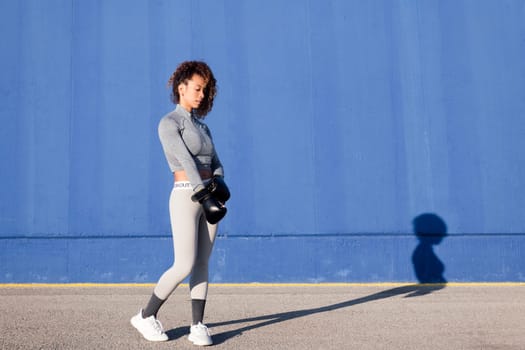 determined adult female boxer putting on boxing gloves during workout on blue background, concept of strength and healthy lifestyle, copy space for text