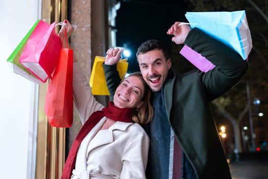 A cheerful couple enjoys a night of shopping, holding up colorful bags with excitement. They are dressed warmly and share smiles, celebrating their successful shopping spree in a vibrant city setting.