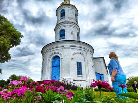 A woman gazes upward at an iconic white tower church with blue accents, framed by colorful flowers on a cloudy day.