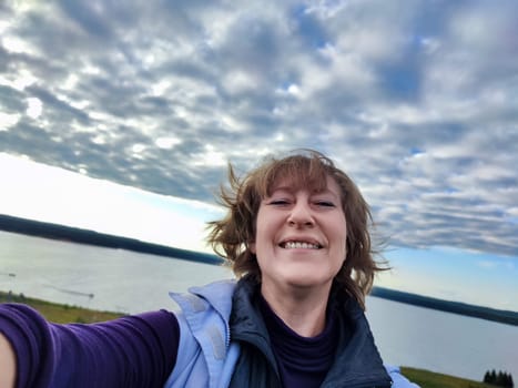 A woman smiles brightly while taking a selfie by the lake, surrounded by stunning clouds and the serenity of nature nearby.