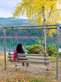 A woman wearing a red scarf sits on a swing type bench enjoying the autumn colors and the river in eastern Washington.