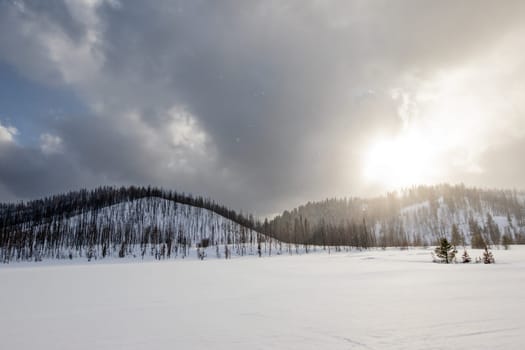 A serene snowy mountain landscape under a dramatic cloudy sky during sunset. The sunlight peeks through, creating a peaceful and atmospheric winter scene.