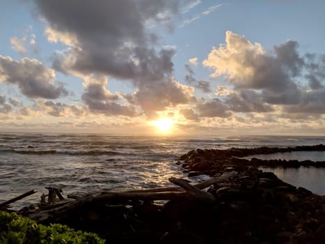 A vibrant sunset illuminates the ocean as colorful clouds drift across the sky, casting a warm glow over the calm waves and rocky shoreline at Lydgate Beach Park.