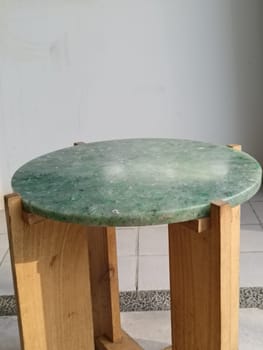 A close-up of a round green marble-topped chair with wooden legs placed on a tiled floor in a bright room.