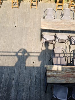 Top view of empty rustic wooden tables with chairs on an outdoor wooden deck, creating a serene and inviting atmosphere. There is also shadows of people from a rooftop