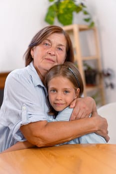 Loving grandmother and granddaughter share warm embrace at home, their faces filled with joy as they look at camera, capturing heartfelt moment of connection across generations