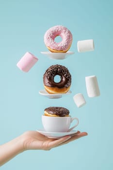 Hand is holding a cup catching flying donuts, chocolate bars and marshmallows on a blue background. This image evokes the concept of sweet breakfast