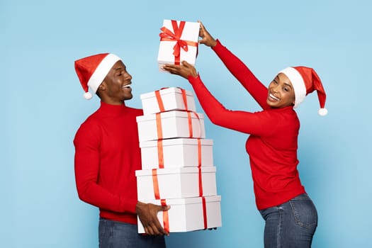 A black couple wearing red sweaters and Santa hats joyfully presents a tall stack of gift boxes wrapped in red ribbons. Their cheerful expressions enhance the festive spirit of the moment.