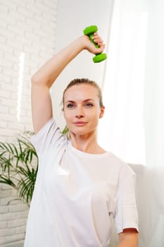 A woman exercises with green dumbbells at home in a bright room filled with light and plants