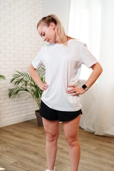 A woman stretches her neck and shoulders in a bright room surrounded by indoor plants during a home workout