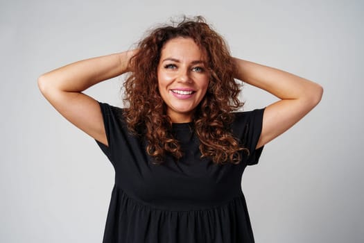 Smiling woman with curly hair poses confidently against a plain background while holding her hands behind her head indoors