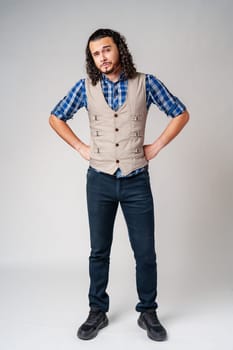 Confident man with curly hair wearing a plaid shirt and vest poses against a neutral background in a thoughtful expression