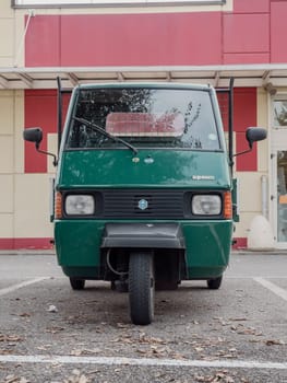 Arquato, PC, italy - October 31st 2024 Front view of a green ape piaggio porter van cargo scooter pickup truck parked in front of a red and beige building, showcasing its unique three wheeled design