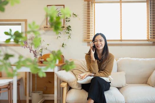 Beautiful young Asian woman talking on mobile phone in her living room.
