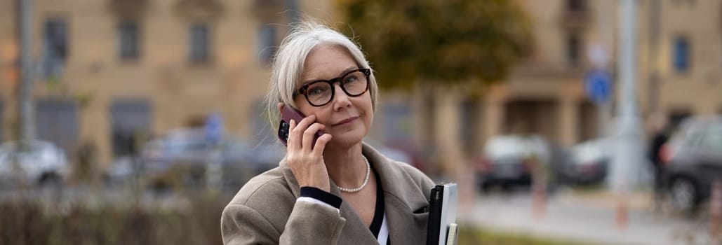 A senior businesswoman stands outside her office, engaged in a phone conversation. She carries a folder and wears glasses, exuding professionalism amidst an urban environment.