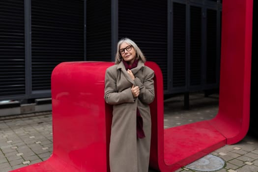 A middle-aged businesswoman stands with a relaxed demeanor, wearing a beige coat and glasses. The striking red architectural element contrasts with her attire, adding vibrancy to the urban landscape.