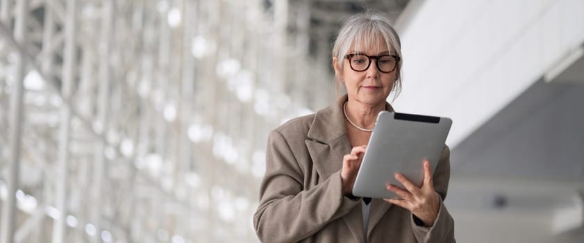 A middle-aged businesswoman is focused on her tablet, engaged in work in a bright, contemporary office setting illuminated by natural light.