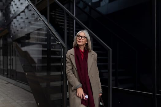 A confident businesswoman in a stylish coat and scarf stands next to sleek, modern stairs, looking thoughtfully into the distance amidst an urban backdrop.