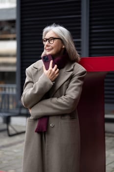 A senior businesswoman stands with a relaxed demeanor outside, dressed warmly in a coat and scarf. She holds a smartphone to her chest, enjoying the cool air.