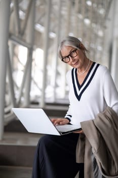 An older woman, dressed in a stylish sweater, is focused on her laptop in a bright, contemporary environment. She holds a coat and looks engaged in her task, enjoying the ambiance.