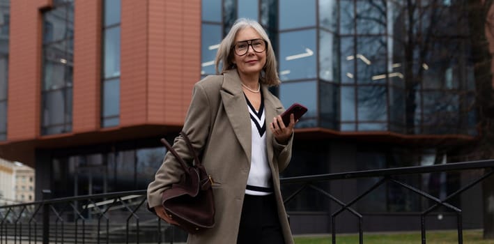A middle-aged businesswoman with gray hair confidently strolls past a contemporary office building, holding her smartphone and wearing a stylish coat and glasses.