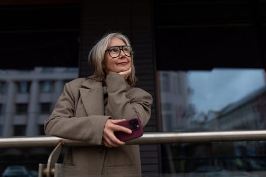 An elderly businesswoman stands thoughtfully with her smartphone, resting her chin on her hand. She is near her office in a vibrant urban area, showcasing a moment of reflection.