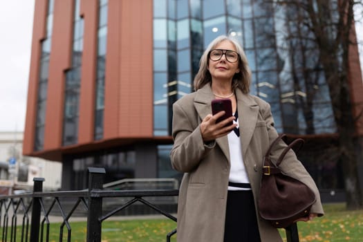 An elderly businesswoman stands confidently outside a contemporary office building, holding her smartphone and wearing a stylish coat and glasses on a brisk day.