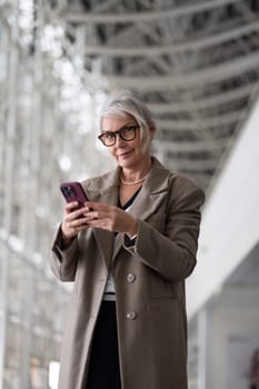 An elderly businesswoman stands confidently in a contemporary office setting, engaging with her smartphone while dressed in a stylish coat. Her expression is focused and professional.