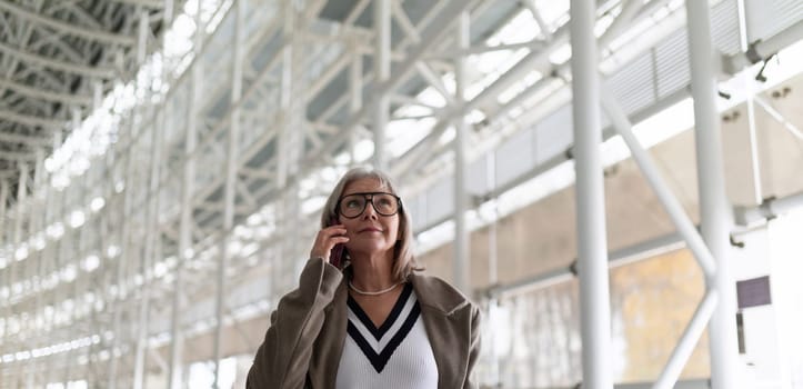 A middle-aged businesswoman with glasses is engaged in a phone conversation while strolling through a bright and spacious airport terminal. Her confident demeanor suggests professionalism and purpose.