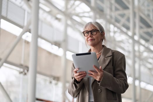 An elderly businesswoman stands outside her office building, holding a tablet and looking poised. She exudes confidence as she prepares for an upcoming meeting.