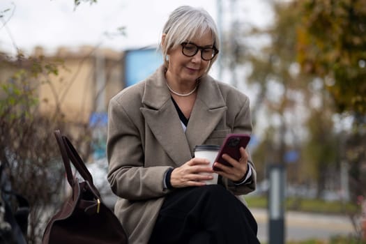 An older woman dressed in a stylish coat and glasses sits on a bench, sipping coffee from a cup while focused on her smartphone in a lively park setting.