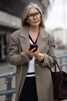 An elderly businesswoman stands outside her office building, dressed in a stylish coat and holding her smartphone, looking thoughtfully into the distance on a cloudy day.