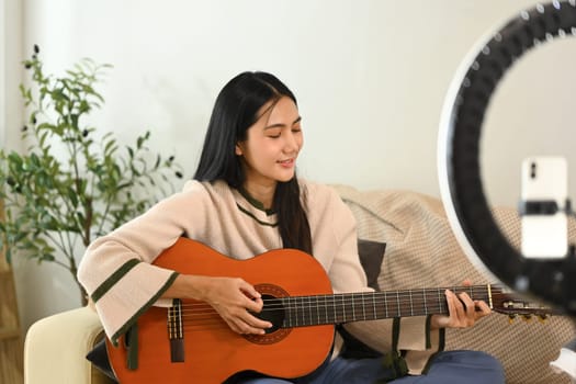 Attractive young woman playing guitar while recording content for social media.