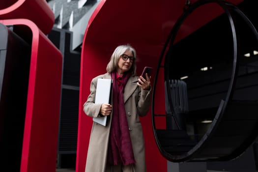 An older woman dressed in a trench coat and scarf stands near vibrant modern structures, intently checking her smartphone while holding a file.