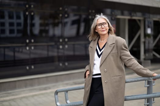 An older woman with gray hair and glasses exudes confidence while leaning against a railing in front of a sleek modern building. She wears a chic outfit that showcases her elegant style.