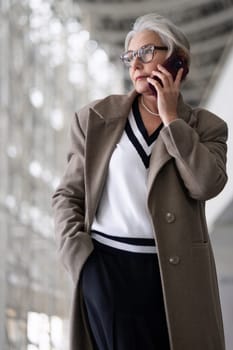 A middle-aged businesswoman wearing an elegant coat engages in a phone conversation while standing in a contemporary office environment filled with natural light.