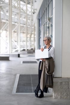 The elderly businesswoman leans against a wall, holding a laptop. She appears engaged in thought, dressed in a stylish outfit that reflects her professional demeanor.