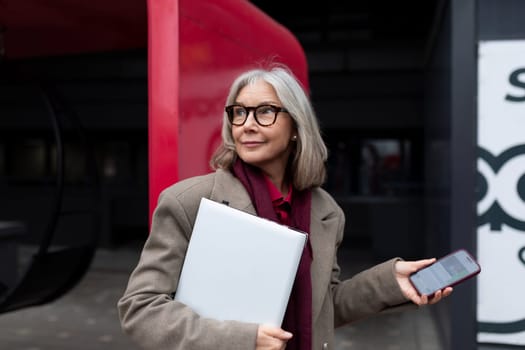 An older woman, dressed in a beige coat and scarf, stands outside a contemporary building, looking thoughtfully to the side as she holds a laptop in one hand and a smartphone in the other.
