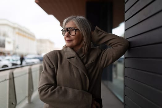The older woman stands confidently with her hand behind her head, dressed in a fashionable coat, observing her surroundings on a cloudy city day.