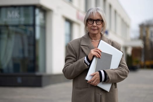 An elderly businesswoman confidently holds a portfolio close to her chest, dressed in a stylish coat while standing outside a contemporary office building on a cool day.