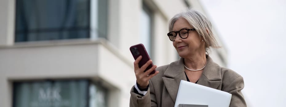 An older woman stands outside an office building, engaged with her smartphone while holding a laptop. She appears focused and relaxed, showcasing modern work-life balance.