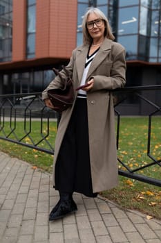 An elderly businesswoman in a fashionable coat poses outside her office building. She holds a bag and looks poised amidst colorful autumn leaves scattered on the ground.