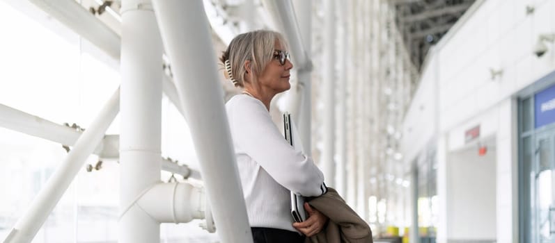 A middle-aged businesswoman stands confidently in a sleek office, reviewing important documents. She appears focused and engaged in a professional setting.