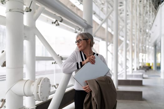 An older woman with glasses stands relaxed in a contemporary architectural space, holding a laptop and a coat. The sunlight filters through large windows, creating a warm atmosphere.