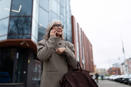 A middle-aged businesswoman dressed in a stylish coat stands on the street, engaged in a phone conversation while holding her handbag. The urban setting features modern buildings.