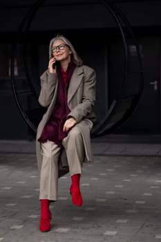 An elderly businesswoman sits on a circular swing outside her office, engaged in a phone conversation. She is dressed in professional attire, showcasing her unique style with bright red socks.