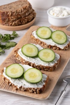 Three slices of nutritious whole grain bread are spread with creamy cheese and topped with fresh cucumber rounds, arranged on a wooden board for a light snack.
