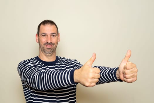 Bearded hispanic man in his 40s wearing a striped sweater with his arms outstretched and thumbs up in a sign of approval and positivity, isolated on a beige background